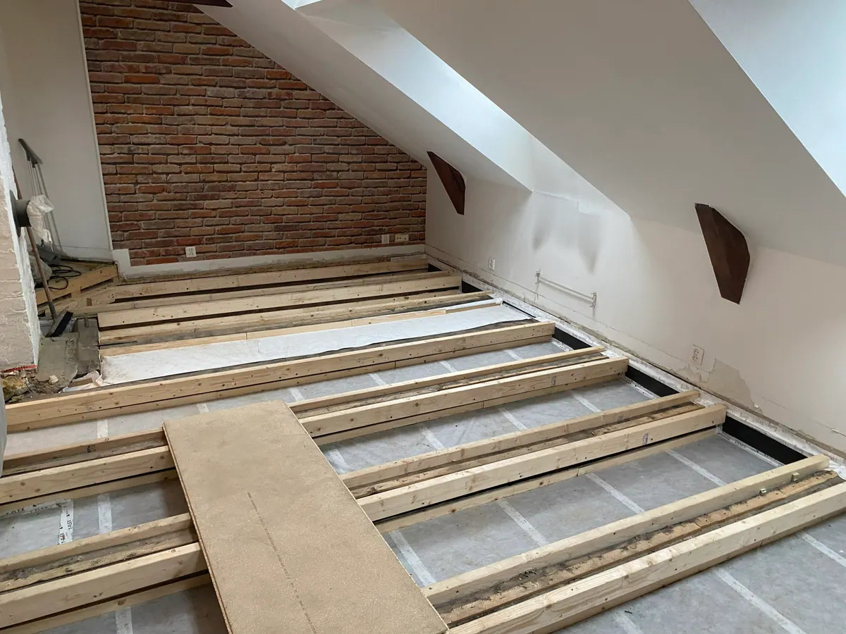 Åpnet loftsgulv med synlige bjelker og teglvegg under arbeid
